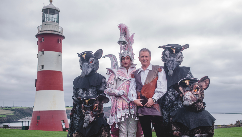 Group of performers in elaborate costumes, including three dressed as large rats and two in ornate outfits, standing on grass near Smeaton’s Tower lighthouse with a cloudy sky in the background.